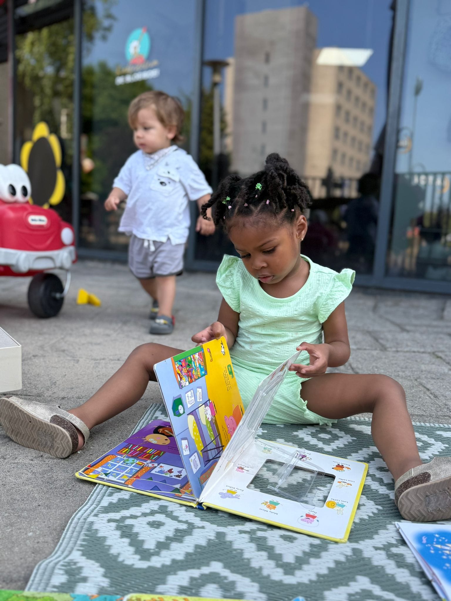 Op een zonnige dag zit een kind op het schoolplein en bekijkt een vrolijk kinderboekje bij Kindercentrum De Bloeiboei.