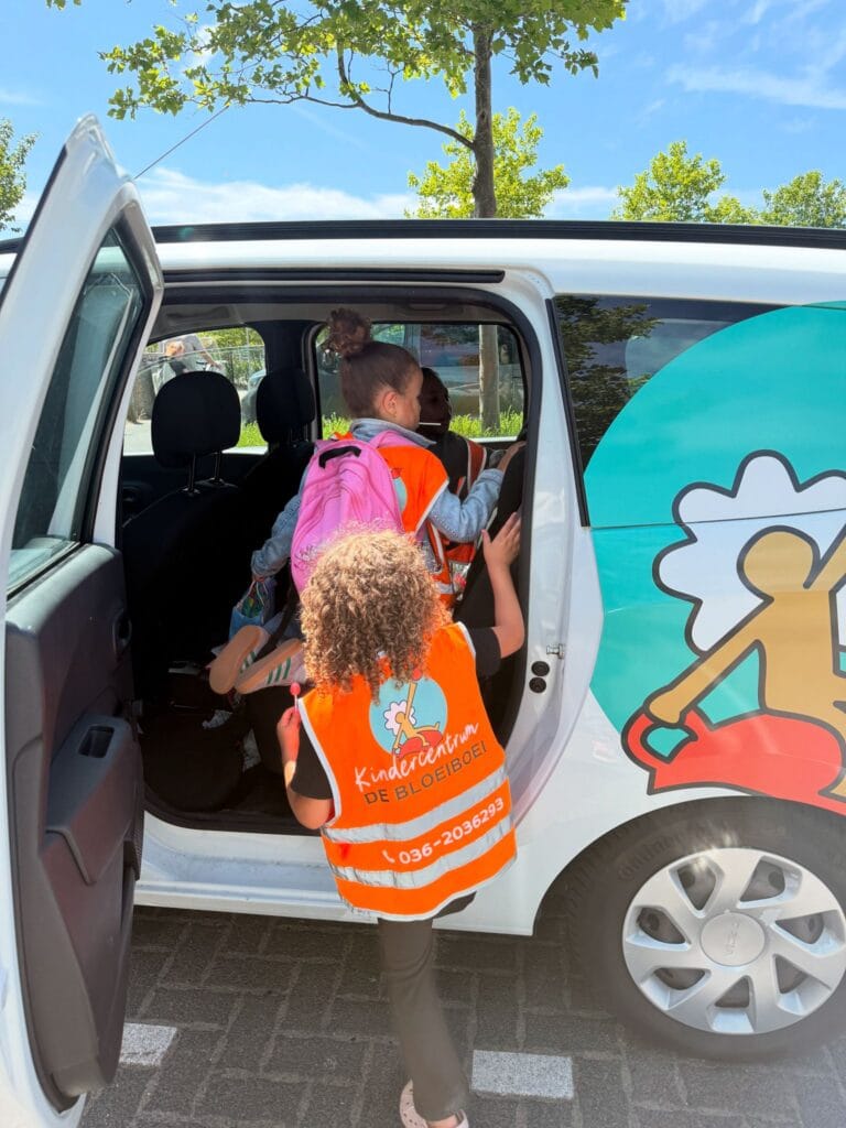 Twee kinderen dragen oranje hesjes met het logo van Kindercentrum De Bloeiboei terwijl ze de auto instappen.